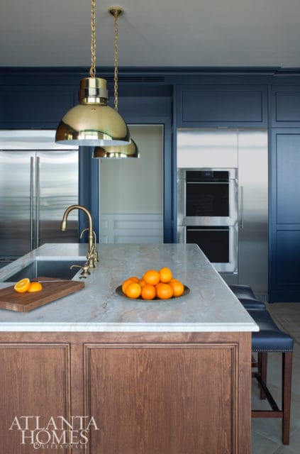 Navy blue kitchen island with marble countertop, brass faucet, and wood paneled base. Stainless steel appliances and pendant lights are visible.