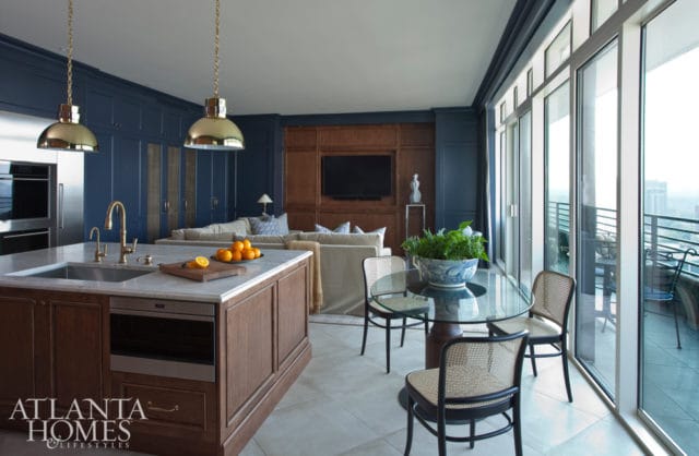 Open concept living area with a dark blue paneled wall, a wood-paneled entertainment center, and a large island with a marble countertop. A glass-top dining table with cane chairs sits near a balcony with city views.