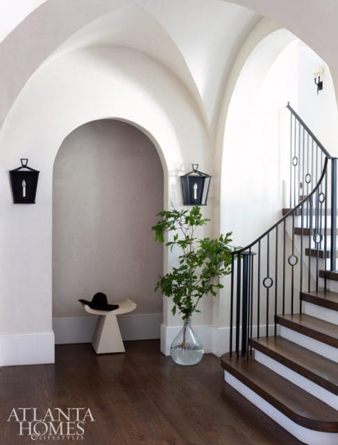Foyer with arched doorways, dark wood floors, a white sculptural stool with a black hat, a large glass vase with greenery, and a dark metal spiral staircase with wooden treads.