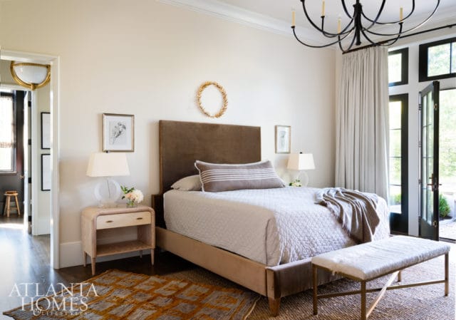 Bedroom with a brown velvet headboard, white quilted bedding, and a textured rug. A gold wreath hangs above the bed, and French doors open to an exterior view.