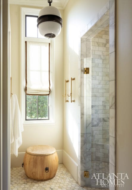 Cream bathroom with marble subway tile shower, hexagonal floor tiles, a wooden drum stool, and brass accents.
