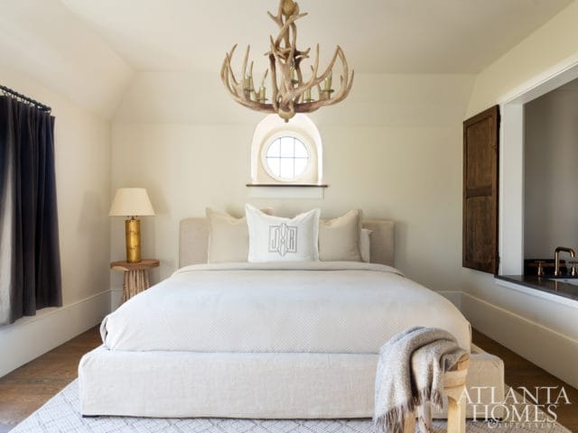 Bedroom with a king-sized bed, neutral linens, a large antler chandelier, and a round window with wooden shutters.