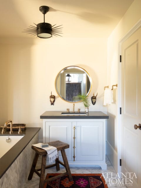 Bathroom vanity with white cabinets, dark countertop, gold hardware, and round mirror. A wooden stool sits in front of the vanity with a small rug on the floor.