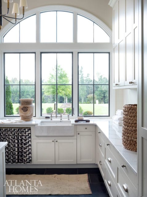 White laundry room with a large farmhouse sink under a wall of windows overlooking a green lawn. White cabinetry with brass hardware lines the counter.