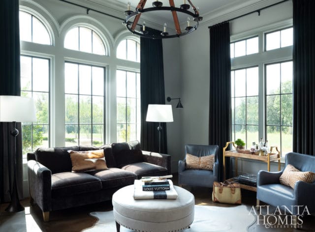 Living room with dark velvet sofa, blue leather armchairs, and large arched windows. A round ottoman sits in front of the sofa.