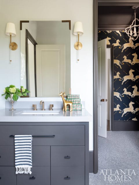 Gray vanity with white countertop and brass faucet, flanked by brass sconces. A doorway reveals a room with zebra wallpaper and a chandelier.