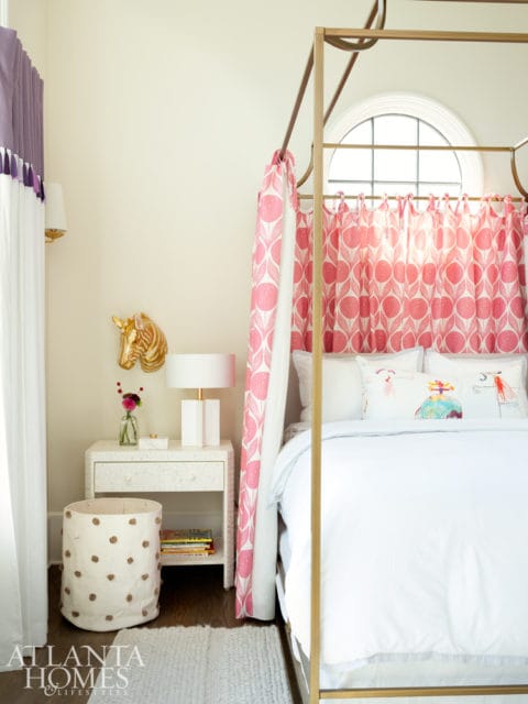 Children's bedroom with a gold canopy bed, pink floral curtains, white nightstand with a gold horse head decor, and a white storage bin with brown polka dots.
