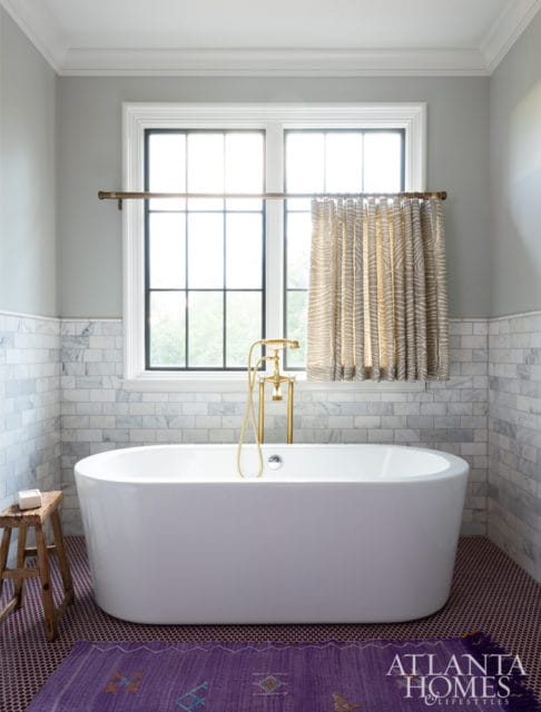 Freestanding white bathtub with brass fixtures, marble tile walls, and a patterned rug on a mosaic tile floor. A small wooden stool sits to the left.