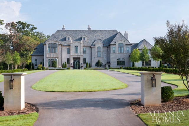 Large stone mansion with gray shingle roof, arched windows, and manicured lawn, with stone pillars and lanterns flanking the driveway.