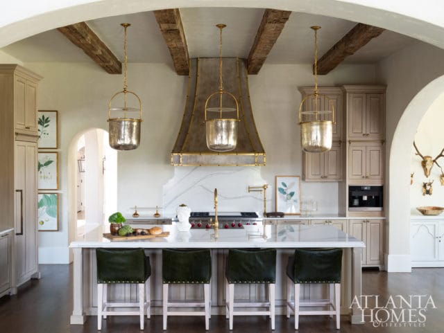 Kitchen with light wood cabinetry, a large metal range hood with marble backsplash, and three glass pendant lights. A white island with dark green bar stools sits in the foreground.