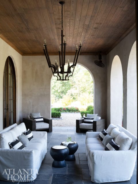 Covered porch with white slipcovered sofas, dark wood chairs, black accent tables, and a wrought iron chandelier under a wood plank ceiling.