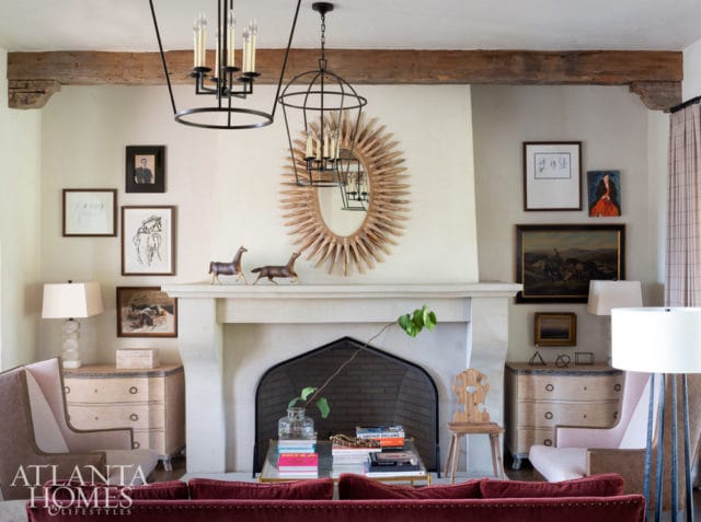Living room with a stone fireplace, sunburst mirror, two chandeliers, and artwork above the mantel. Two chairs and a sofa are visible in the foreground.