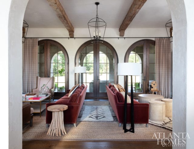 Living room with two red velvet sofas, arched French doors, exposed wooden beams, and a rustic driftwood side table.