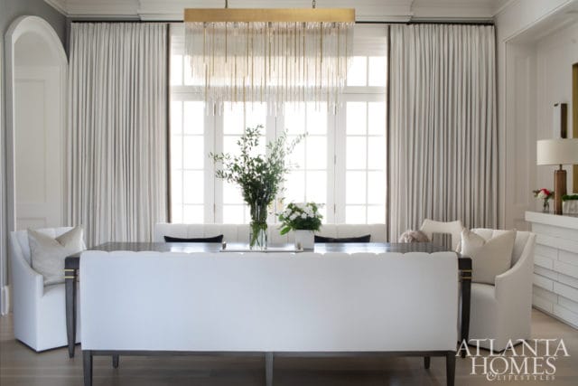 Dining room with a dark wood table, white upholstered chairs and banquette seating, a crystal chandelier, and floor-to-ceiling white curtains.
