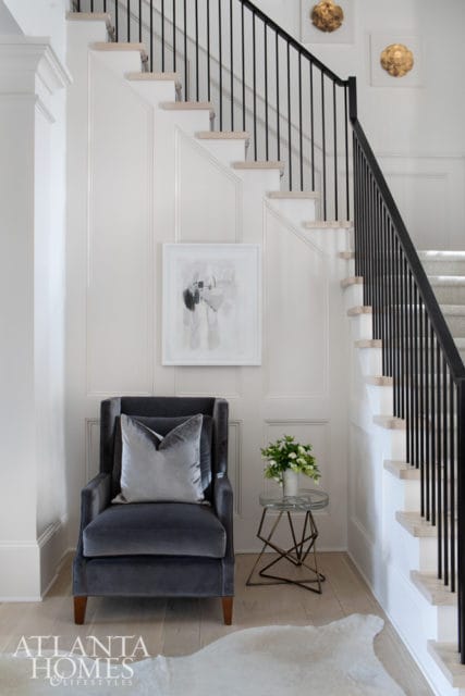 Gray velvet armchair with a silver pillow in a hallway with paneled walls, a modern staircase with black railings, and abstract art.