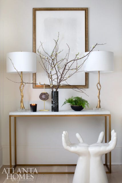Marble console table with gold frame topped with two white lamps, a vase of branches, a fern, and a black candle. A white hand-shaped stool sits in front.