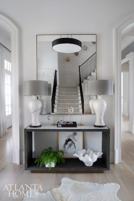 Foyer console table with two white lamps, marble top, and framed art. Mirror reflects a staircase with black railing and a large drum pendant light.