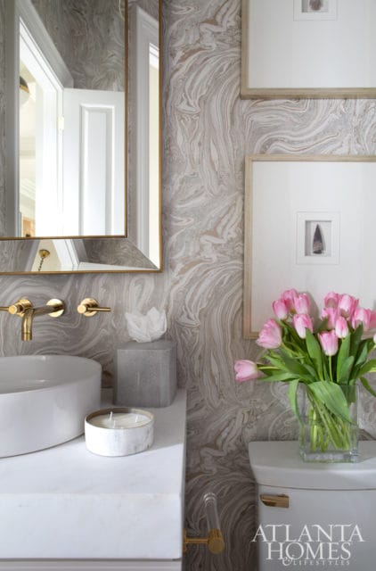 Powder room with swirled marble wallpaper, white countertop with vessel sink and gold faucet, and a vase of pink tulips on the toilet tank.
