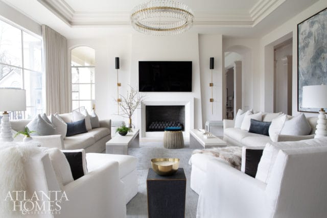 Bright living room with white sofas, grey accent pillows, a white fireplace with a black television above, and a large circular crystal chandelier.