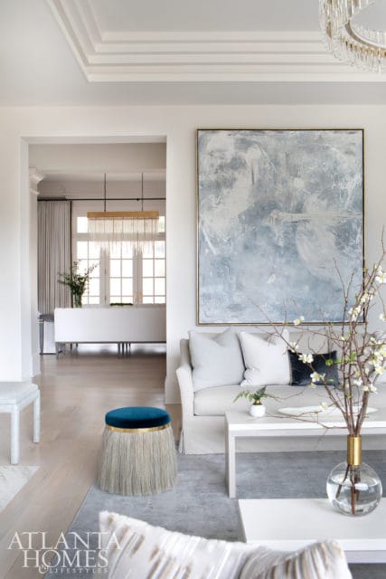 Living room with a light gray sofa, abstract blue and white artwork, and a teal ottoman with fringe. A view into a dining room with a modern chandelier is visible through an archway.