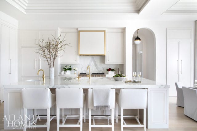 White kitchen with marble island and countertops, brass accents on range hood and faucets, and white bar stools.