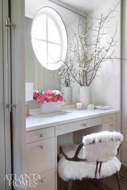 White vanity with a round window above, a white vase with branches and white flowers, a white vase with pink and white peonies, and a white fur-covered chair.