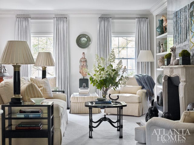 Living room with neutral-colored furniture, including sofas and armchairs, a fireplace with decorative objects, and tall windows with striped curtains.