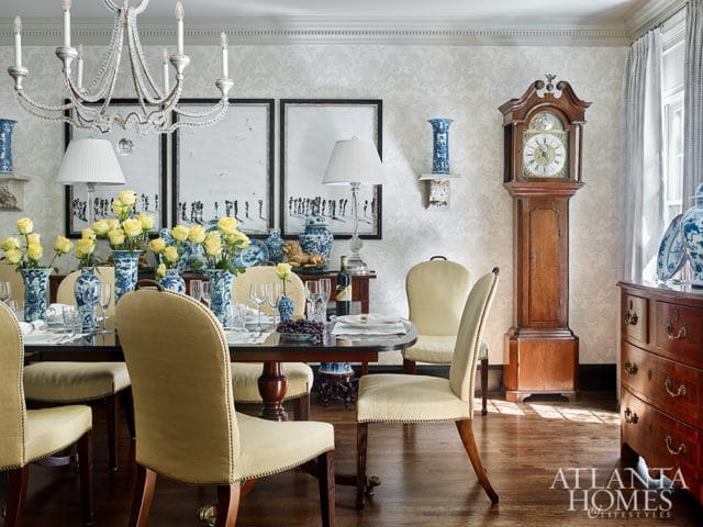 Dining room with a dark wood table set for a meal, surrounded by upholstered chairs. Yellow roses in blue and white vases adorn the table. A grandfather clock stands to the right, with a patterned wallpaper and ornate chandelier overhead.