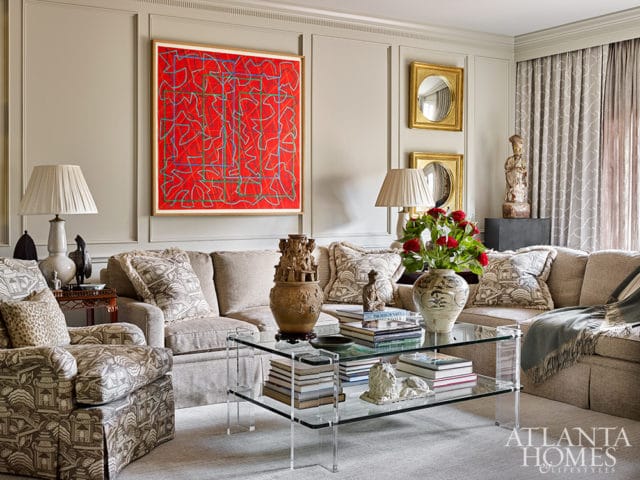 Living room with neutral-toned sofas and armchairs, a red abstract artwork, a glass coffee table, and patterned curtains.