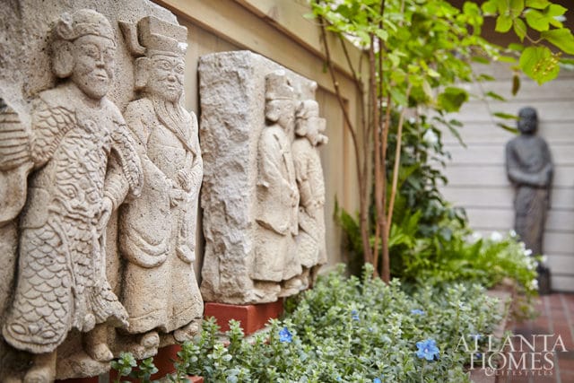 Stone relief carvings of figures in traditional dress stand in a garden with lush greenery, small blue flowers, and a distant statue.