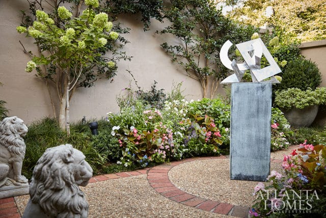 Sculptural garden with gravel pathway, brick edging, flowering plants, stone lion statues, and a modern metal sculpture on a pedestal.