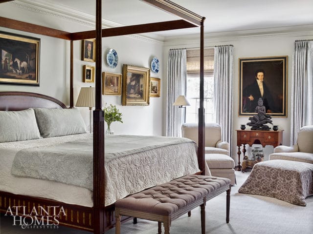 Bedroom with a dark wood four-poster bed, light gray bedding, and a tufted bench. Walls are adorned with framed paintings and blue and white plates.
