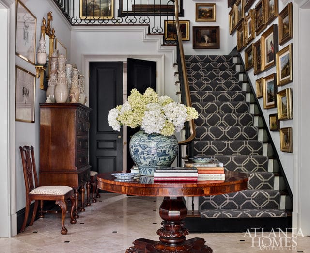 Foyer with a dark wood chest, round table with books and a vase of hydrangeas, patterned carpeted stairs, and walls adorned with framed art.