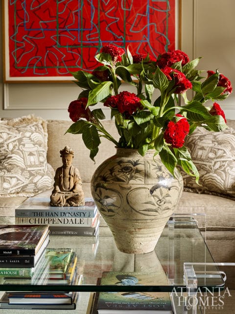 Living room vignette with a large ceramic vase of red flowers on a glass coffee table, a small Buddha statue, stacked books, and abstract red art on the wall.