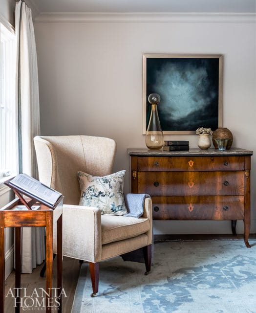 Wingback armchair with patterned pillow and blue throw next to a wooden dresser with marble top and abstract art above.