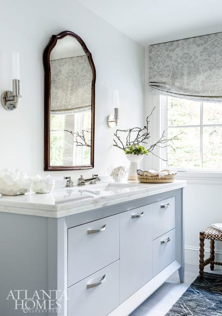 Light blue bathroom vanity with marble countertop, brushed nickel hardware, and a decorative mirror above. White walls with sconces and a patterned Roman shade window treatment.