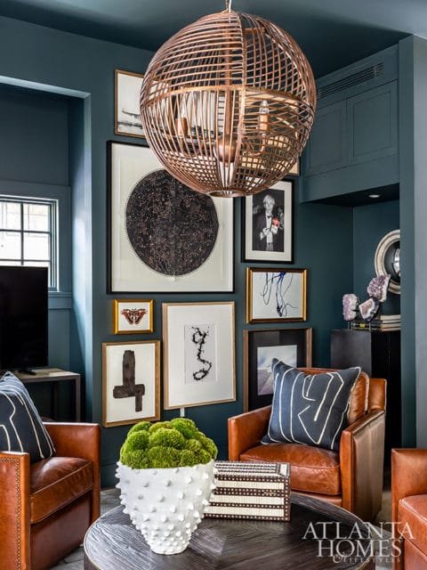 Dark teal living room with leather armchairs, gallery wall featuring a constellation map and abstract art, a large copper pendant light, and a white textured vase filled with moss.