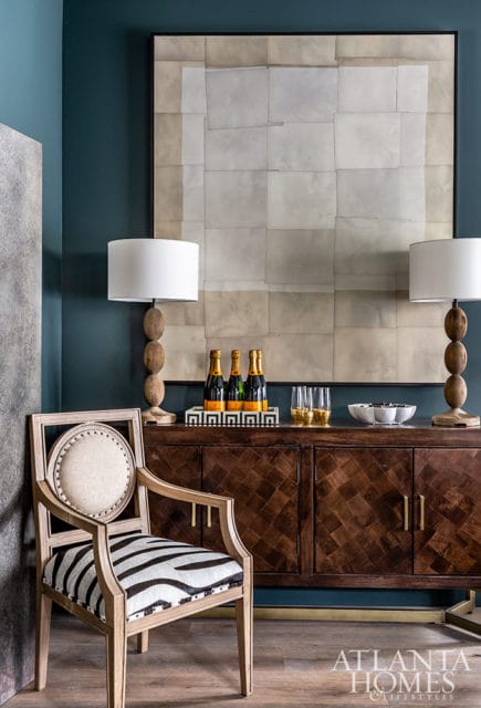 Living room with teal walls, a wooden sideboard with a geometric pattern, two lamps, a framed abstract art piece, a chair with zebra print upholstery, and a tray of champagne bottles.