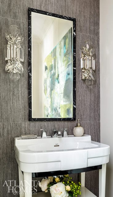 Modern bathroom with a white sink, black marble framed mirror reflecting abstract art, and crystal sconces on textured gray wallpaper.