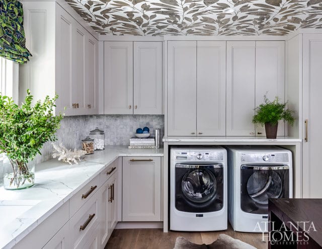 Laundry room with light gray cabinetry, marble countertops, and a patterned leaf wallpaper on the ceiling. Two white washing machines are visible below the counter.