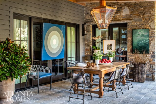 Covered patio with a wooden dining table and chairs, a large blue abstract painting, and stone walls.