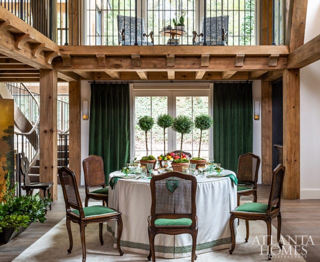 Dining room with a round table set for a meal, surrounded by cane-backed chairs with green upholstery. Exposed wooden beams and a spiral staircase are visible.