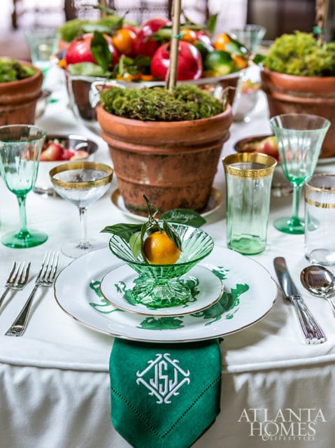 Table setting with white plates, green glassware, and a green napkin with monogrammed initials. A small orange sits in a green glass dish.