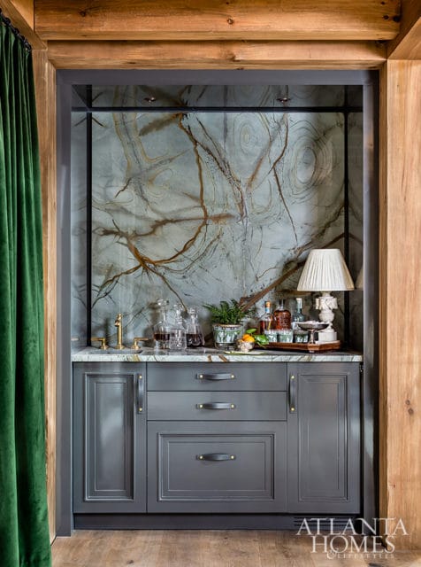 Gray built-in bar with marble countertop, brass faucet, and dramatic veined wallpaper. Features decanters, bottles, a lamp, and green velvet curtains.