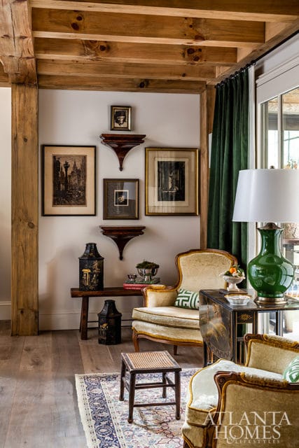 Living room with exposed wooden beams, a collection of framed art and decorative objects on the wall, a green velvet curtain, and upholstered chairs with ornate detailing.