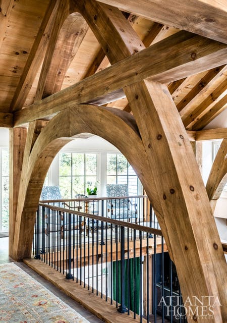 Interior view of a home with exposed wooden beams, an arched wooden entryway, and a black metal railing overlooking a lower level.