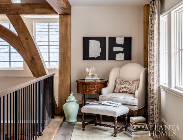 Cream upholstered armchair with patterned pillow, ottoman, and side table with books and decor, next to a large mint green vase and patterned curtains. Exposed wooden beams and a railing are visible.