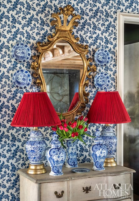 Entryway console table with blue and white ginger jars, red pleated lampshades, and a gold ornate mirror against blue floral wallpaper.