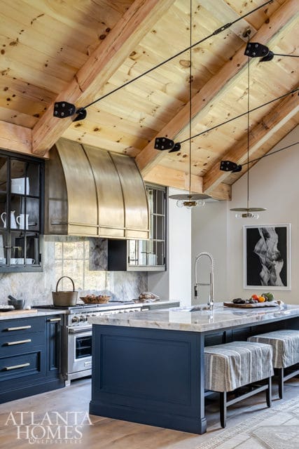 Navy blue kitchen island with marble countertop, flanked by striped stools. Features a stainless steel range with a brass hood, marble backsplash, and wood-beamed vaulted ceiling with industrial pendant lights.