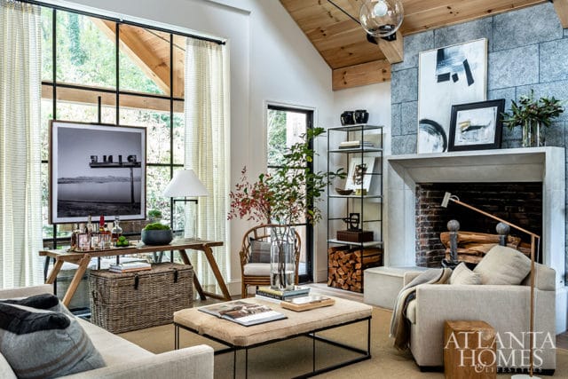 Living room with a large stone fireplace, wood-beamed ceiling, and a large window overlooking greenery. Furnishings include a neutral sofa, armchair, and a coffee table ottoman.